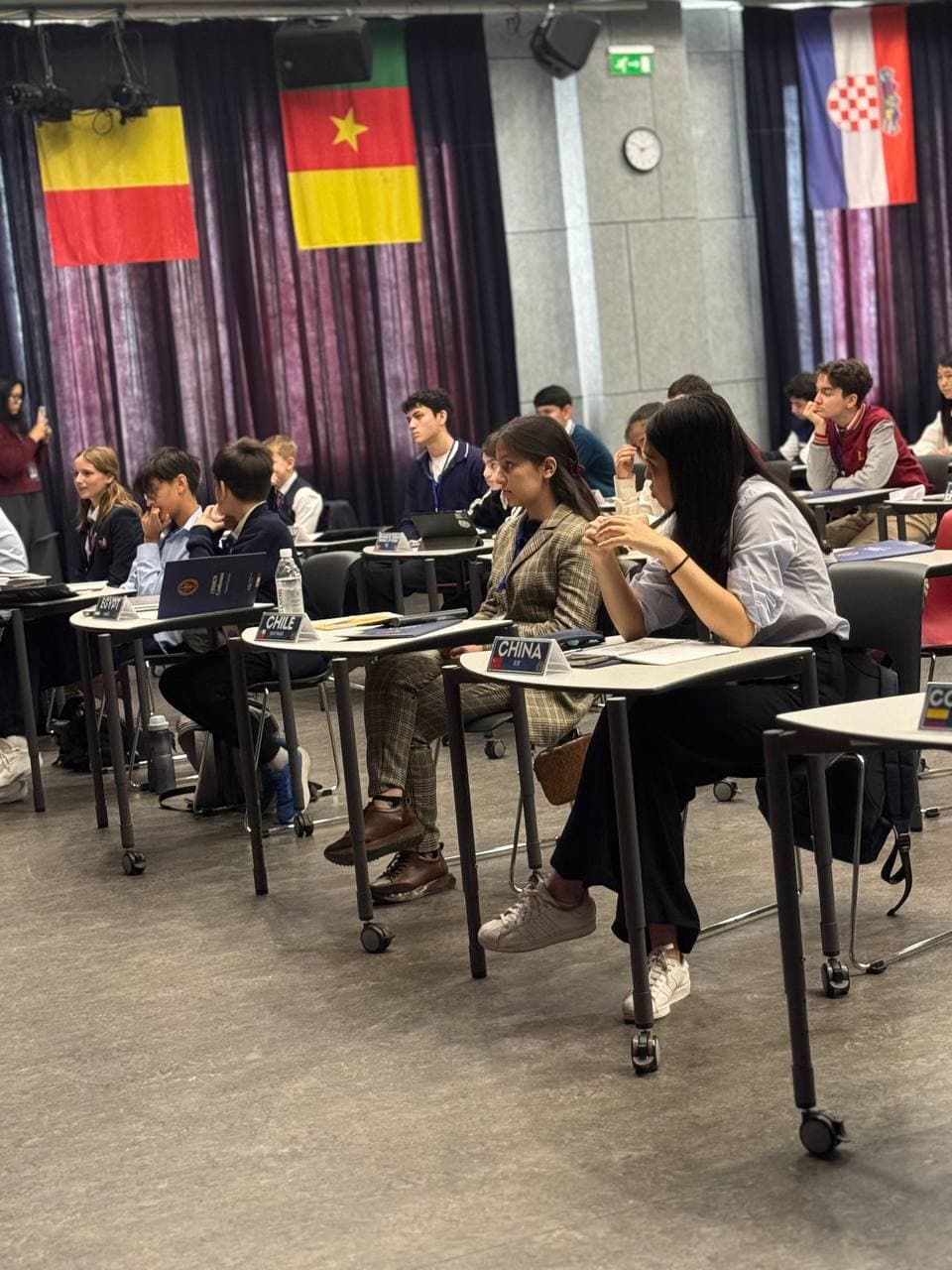Katta zalda o'tkazilgan MUN majlisi, sahna orqasida ishtirokchi davlatlar bayroqlari